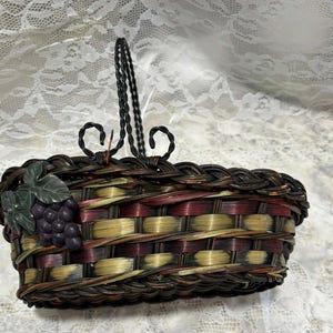 May include: Woven basket with a decorative handle and grape cluster accent. The basket features a mix of brown, red, and yellow tones. The handle is black and has a decorative swirl design. The basket is likely for decorative use.