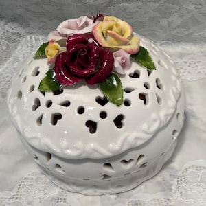 May include: A white ceramic trinket box with heart-shaped cutouts and a lid adorned with colorful ceramic roses and green leaves. The roses are in shades of red, pink, and yellow. The box has a scalloped edge.