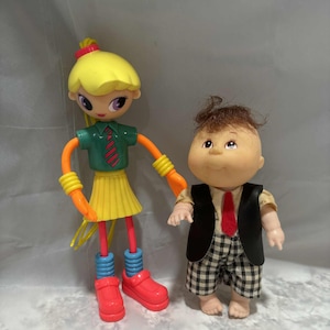 May include: Two vintage dolls. One doll has a yellow head, green shirt, yellow skirt, and red shoes. The other doll has brown hair, a black vest, and checkered shorts. Both dolls are standing.