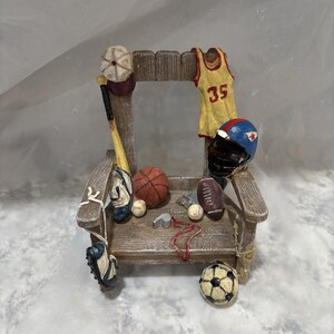 May include: A wooden chair with a variety of sports equipment, including a baseball bat, basketball, football, soccer ball, and a baseball cap. The chair is decorated with a red, white, and blue football helmet and a yellow jersey with the number 35 on it.