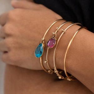 Puede incluir: Brazaletes dorados, algunos adornados con piedras preciosas en forma de lágrima en azul y rosa. Las pulseras están apiladas en una muñeca, mostrando su delicado diseño y variaciones de color. La joyería es simple y elegante.