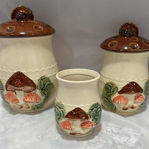 Può includere: Un set di tre barattoli da cucina in ceramica e un piccolo vaso. I barattoli hanno coperchi marroni e sono decorati con un motivo a fungo nei toni del marrone, rosso e verde. Il vaso ha un motivo a fungo simile.
