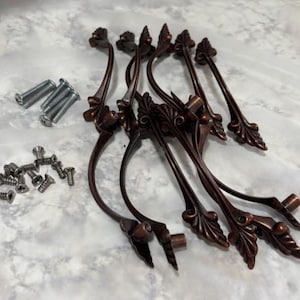 May include: A set of eight bronze-colored cabinet pulls with decorative leaf and scroll details. Includes silver screws and bolts for installation. The pulls are approximately 6 inches long.
