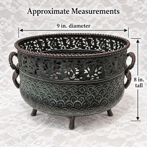 May include: A dark metal bowl with a rope-textured rim, two side handles, and three feet. The upper section features a cut-out floral pattern, while the lower body has a scalloped texture. It measures 9 inches in diameter and 8 inches tall.