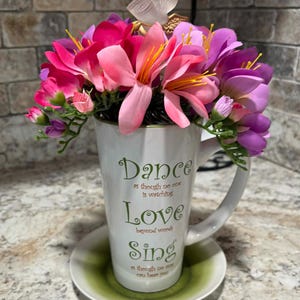 May include: A white ceramic mug with a handle, filled with vibrant pink and purple artificial flowers. The mug has the words "Dance", "Love", and "Sing" printed on it. The mug sits on a green-rimmed saucer, with a decorative angel figurine on top.