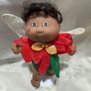 Puede incluir: Una muñeca Cabbage Patch Kid vestida como un hada de Navidad. La muñeca tiene piel morena, cabello negro y lleva un disfraz de poinsettia rojo con alas verdes.
