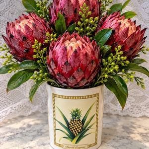 Puede incluir: Un recipiente cilíndrico de cerámica blanca con una ilustración de piña y el texto Smooth Cayenne, lleno de un ramo de cuatro grandes flores de protea de color rojo oscuro, hojas verdes y pequeños racimos de bayas de color verde claro.