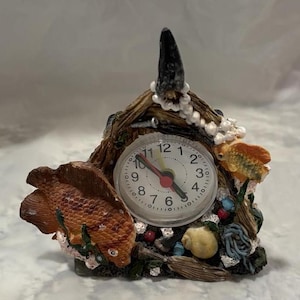 Könnte beinhalten: Eine kleine, dekorative Uhr in Form eines Hauses mit einem Fisch und einem Zifferblatt. Das Zifferblatt ist weiß mit schwarzen Zahlen und Zeigern. Das Haus ist aus braunem und beigefarbenem Material gefertigt und hat eine kleine, weiße Perlenkette darum. Der Fisch ist orange und hat einen weißen Bauch.