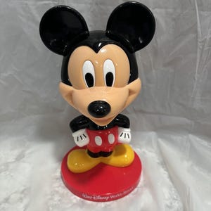 May include: A Mickey Mouse figurine with a black head and ears, a tan face, and a red and white outfit. The figurine stands on a red base with the text "Walt Disney World Resort". The figure has yellow shoes and white gloves.