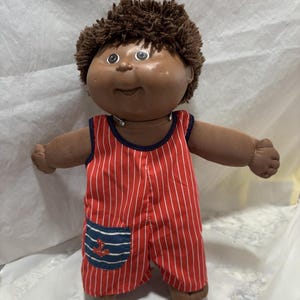 Peut inclure: Une poupée Cabbage Patch Kid brune avec des cheveux bouclés bruns portant une salopette rayée rouge et blanche avec une poche bleue à ancre.