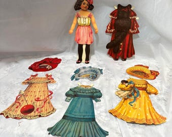 May include: A collection of vintage paper doll clothing and accessories. The doll is wearing a pink dress. The outfits include a red coat, a yellow dress, a blue dress, and a hat. Accessories include a hat and a mask. The items are in a variety of colours.