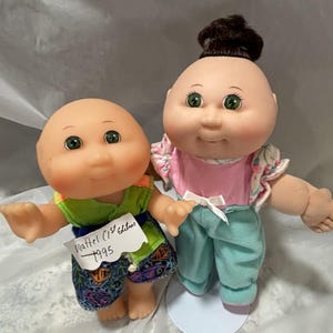 May include: Two Cabbage Patch Kids dolls, one with brown hair and a pink shirt and teal pants, and one with a green shirt and colorful pants. The doll with brown hair has green eyes and a smiling face. The doll with the green shirt has green eyes and a smiling face. The doll with the green shirt has a tag that says "Mattel (C) 1995 Edition".