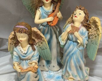 Puede incluir: Tres figuras de ángeles tocando instrumentos musicales. El ángel del centro está tocando una mandolina, el ángel de la izquierda está tocando una flauta dulce y el ángel de la derecha está tocando una flauta. Los ángeles llevan todos vestidos azules y tienen alas.