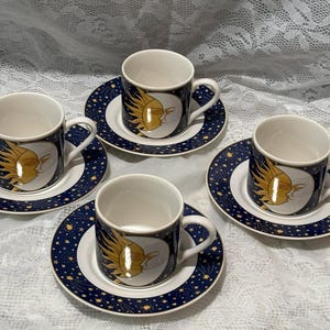 May include: A set of four ceramic espresso cups and matching saucers featuring a dark blue background with a gold sun and moon celestial motif. Each cup has a white interior and a handle, while the saucers display a matching starry night pattern.