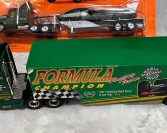 May include: Green and yellow toy semi-truck with a trailer featuring "FORMULA CHAMPION" text and a race car graphic. The truck has a checkered flag design and the word "POWER" on the cab. Another toy truck with a trailer carrying a jet is visible in the background.