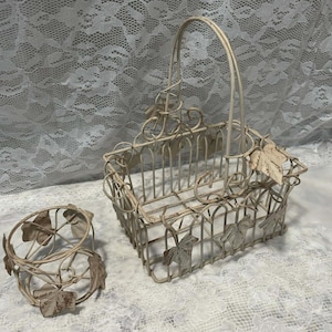 May include: Two off-white metal baskets with leaf accents. One is a rectangular basket with a handle, and the other is a smaller, round basket. Both have a distressed, vintage appearance.