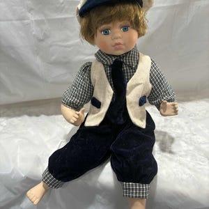 Puede incluir: Muñeca de porcelana de un niño, con una gorra color crema con borde azul marino. Tiene ojos azules y cabello castaño claro. La muñeca viste una camisa a cuadros, un chaleco color crema, una corbata azul marino y pantalones de terciopelo azul marino.