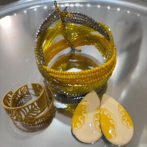 Könnte beinhalten: Eine Auswahl an Schmuck, darunter ein Armband aus gelben und silbernen Perlen, ein Paar gelbe, blattförmige Creolen und zwei tropfenförmige Ohrringe mit gelben und weißen Designs.