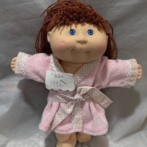 May include: A Cabbage Patch Kid doll with brown yarn hair wearing a pink robe with white polka dots and a white lace trim. The doll has blue eyes and rosy cheeks. A white label on the robe says "By Hasbro 1992".