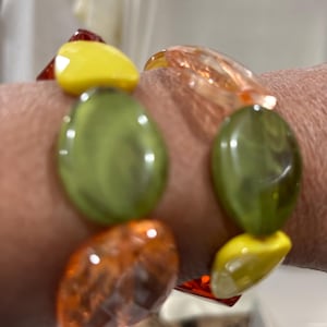 Könnte beinhalten: Ein farbenfrohes Armband mit großen, unregelmäßig geformten Perlen. Die Perlen sind in Gelb-, Grün- und Orangetönen gehalten und haben ein facettiertes, durchscheinendes Aussehen. Das Armband wird am Handgelenk getragen.