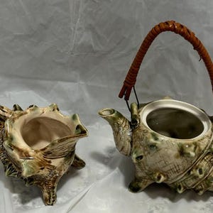 May include: A ceramic teapot and creamer set shaped like seashells. The teapot has a brown leather handle and a lid. The creamer is a smaller seashell with a spout.