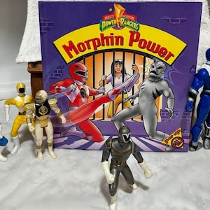 May include: A collection of Power Rangers action figures, including the Red Ranger, Yellow Ranger, White Ranger, and Blue Ranger. The figures are posed in action stances and are standing in front of a book titled "Mighty Morphin Power Rangers: Morphin Power".