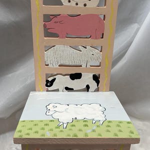 May include: A pink wooden children's chair with a yellow stripe down the side. The back of the chair is decorated with painted farm animals, including a chicken, pig, sheep, and cow. The seat of the chair is painted with a green field and a sheep.