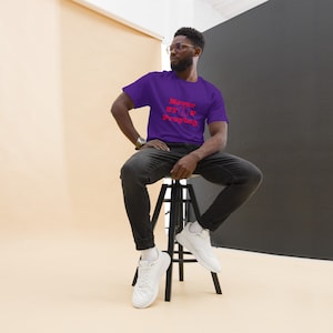 Puede incluir: Un hombre con una camiseta morada con el texto "Never Stop Praying" en letras rojas. Está sentado en un taburete negro y lleva pantalones vaqueros negros y zapatillas blancas.