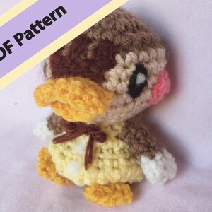 May include: A crocheted stuffed animal resembling a duck with brown, yellow, and white yarn. The duck has a pink blush on its cheek and a brown bow around its neck.