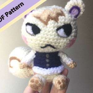 Puede incluir: Un juguete de peluche tejido a crochet de un animal blanco con pelo marrón en la cabeza, que lleva un chaleco azul y una falda blanca. El juguete tiene orejas moradas y mejillas rosadas.