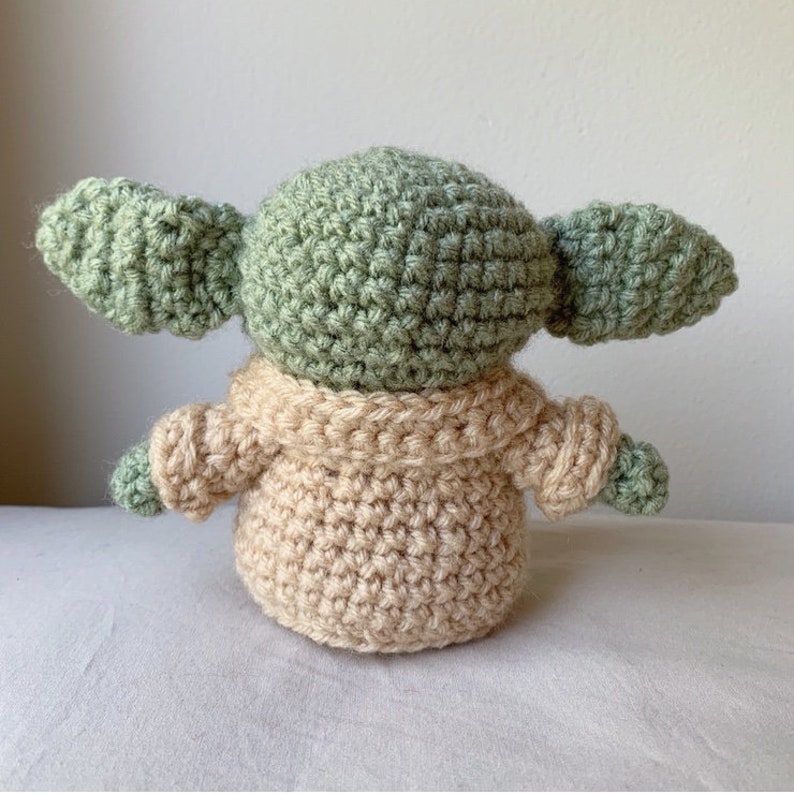 Puede incluir: Un juguete de peluche tejido a crochet con forma de un personaje de la franquicia Star Wars. El juguete es verde y tiene orejas grandes. Lleva un su&eacute;ter tejido a crochet de color beige.