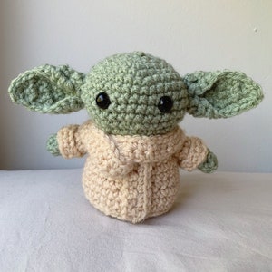Puede incluir: Un juguete de peluche verde y beige tejido a crochet con ojos grandes. El juguete lleva un abrigo tejido a crochet beige.