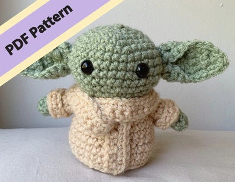 Puede incluir: Un juguete de peluche tejido a crochet de un personaje verde con orejas grandes, que lleva un su&eacute;ter beige. El juguete tiene ojos negros y una peque&ntilde;a boca. La imagen incluye el texto "PDF Pattern".