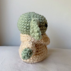 Puede incluir: Un juguete de peluche tejido a crochet de un personaje verde con orejas grandes, que lleva un abrigo beige tejido a crochet. El juguete est&aacute; de pie sobre una superficie blanca.