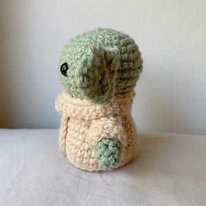 Puede incluir: Un juguete de peluche tejido a crochet de un personaje verde que lleva un abrigo beige tejido a crochet. El personaje tiene ojos negros grandes y una boca peque&ntilde;a.