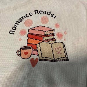 May include: A cross-stitch embroidery on white fabric featuring a stack of books, an open book with a heart, and a mug. The text 'Romance Reader' is stitched in black above the books, surrounded by small pink circular accents and a red heart.