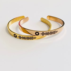 Peut inclure: Deux bracelets de manchette dorés. L'un est doré brillant, l'autre est or rose. Chaque bracelet comporte un symbole de cœur noir et un motif noir en forme de code-barres. Les bracelets sont ouverts et reposent sur une surface blanche.