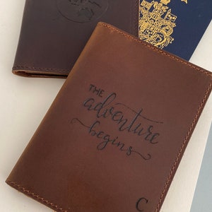 Puede incluir: Dos porta pasaportes de cuero marr&oacute;n. Uno tiene un dise&ntilde;o de globo terr&aacute;queo y el otro tiene el texto "The adventure begins" y las iniciales "C W".