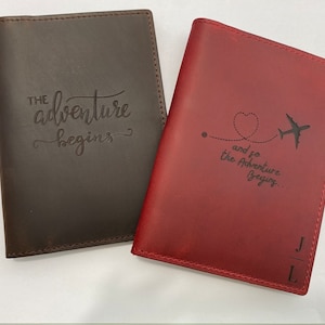 Puede incluir: Dos porta pasaportes de cuero. Uno es marr&oacute;n con el texto "The adventure begins". El otro es rojo con el texto "and so the adventure begins..." y un coraz&oacute;n con un avi&oacute;n volando a trav&eacute;s de &eacute;l.