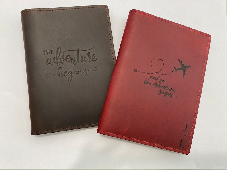 Puede incluir: Dos porta pasaportes de cuero. Uno es marr&oacute;n con el texto "The adventure begins". El otro es rojo con el texto "and so the adventure begins..." y un coraz&oacute;n con un avi&oacute;n volando a trav&eacute;s de &eacute;l.