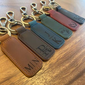 May include: A collection of personalized leather keychains in various colors, including brown, green, and black. Each keychain features engraved initials or names, attached to gold-colored metal hardware.