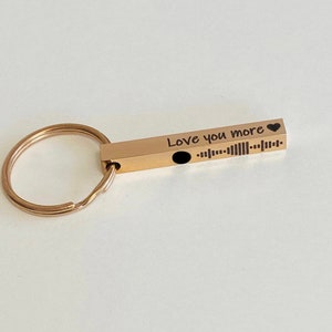 May include: Gold-coloured keychain with a bar engraved with the text "Love you more" and a small heart symbol. The bar also features a black and white barcode.