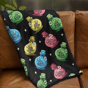 May include: Black throw blanket with a repeating pattern of colourful potion bottles. Each bottle contains a dinosaur skeleton and a tag. The bottles are in shades of blue, green, yellow, and pink, with white star accents.