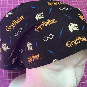 May include: Black scrub cap featuring a repeating pattern of the word "Gryffindor" in gold, white glasses, white lion heads, and blue lightning bolts. The words "Harry Potter" are also visible. The cap is designed to fit a head and is made of fabric.
