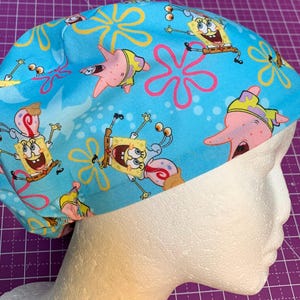 May include: A light blue scrub cap featuring a repeating pattern of SpongeBob SquarePants, Patrick Star, and starfish. The cap is made of fabric and is designed to cover the hair. The background is a light blue color.