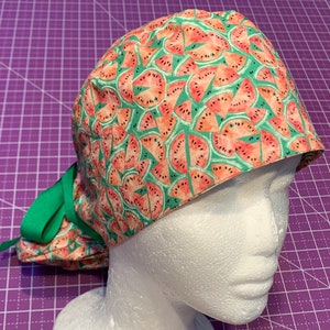 May include: A green and pink surgical scrub hat with a watermelon print. The hat has a tie at the back.