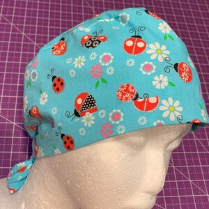 May include: A blue scrub hat with a pattern of ladybugs and white daisies. The hat has a tie at the back.