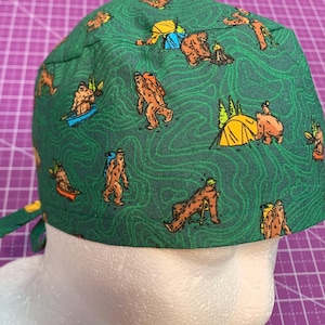 May include: Green scrub cap with a repeating pattern of cartoon Bigfoot figures, tents, and topographic lines. The Bigfoot figures are in various poses, including hiking, kayaking, and camping. The cap has a tie closure.