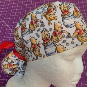 May include: A white scrub cap featuring a repeating pattern of Winnie the Pooh characters in various poses. The cap has a red ribbon tie and is displayed on a mannequin head. The design includes Pooh Bear in a red shirt and blue honey pots.