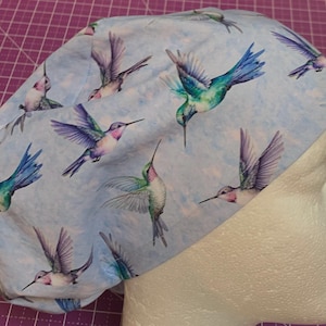 May include: A scrub cap featuring a light blue background with a repeating pattern of colorful hummingbirds in flight. The birds display shades of green, blue, purple, and white, depicted in various poses across the fabric surface of the head covering.